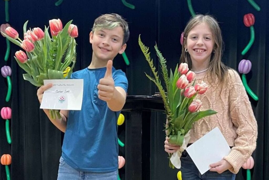 Two young tulip ambassadors stand holding bouquets of tulips and certificates and one of them gives a thumbs up.