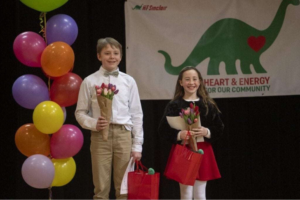 A young boy and a young girl stand on a stage with ballons behind them holding bouquets of colorful tulips.