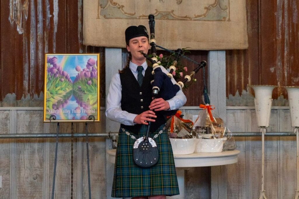 A girl wearing traditional Scottish clothing plays the bagpipes on stage in front of a Skagit Valley Tulip Festival poster